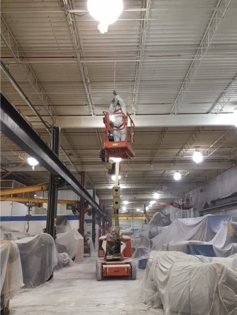 Technician in a lift applying ceiling coating or cleaning overhead; the area below is covered in plastic with equipment wrapped for protection.