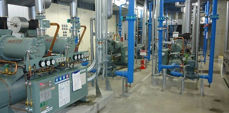 Industrial refrigeration or mechanical room with large compressors, gauges, and blue overhead pipes arranged in rows.