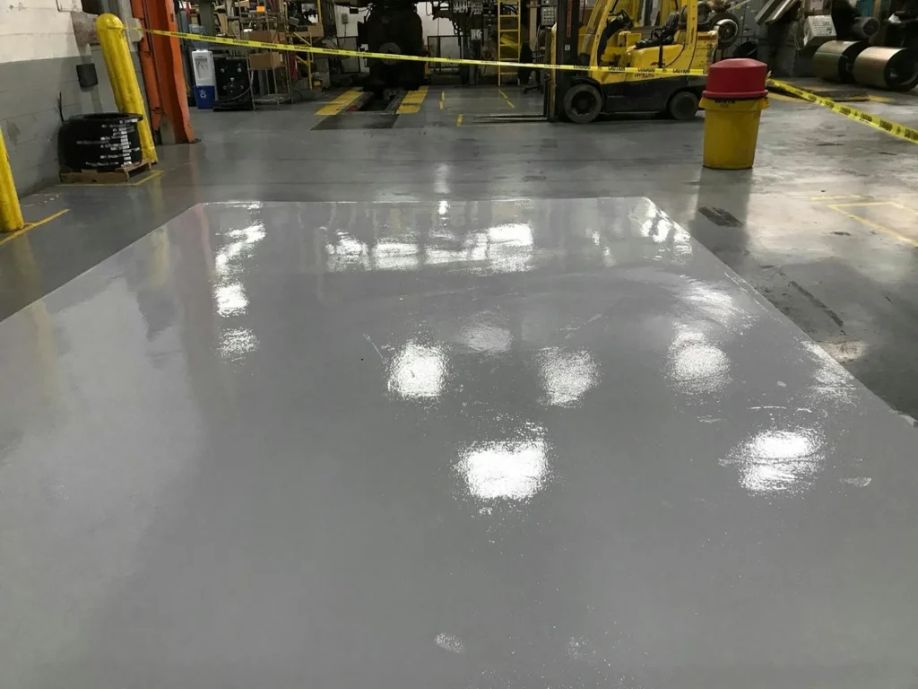 Freshly coated gray epoxy floor patch curing in a production area; the zone is taped off with caution tape, equipment and a forklift in the background.