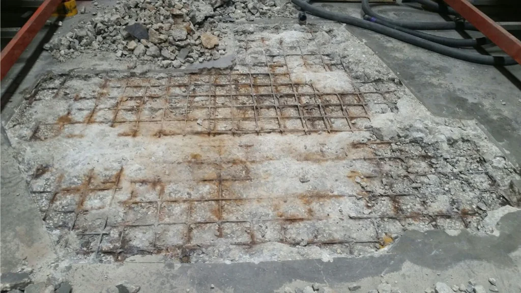 Concrete slab opened to expose a rusted rebar grid; broken rubble and hoses surround the work area.