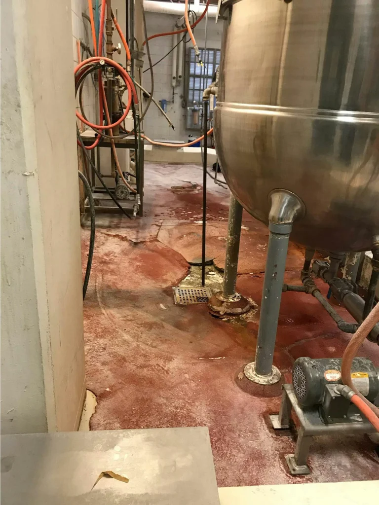 Process room with stainless tank and utility lines above a stained, deteriorated reddish floor prior to repair.