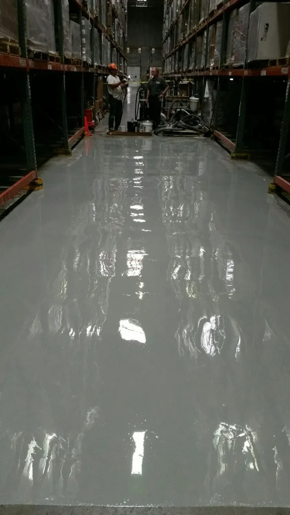 High-gloss gray epoxy coating freshly applied down a warehouse aisle; two workers and equipment at the far end.