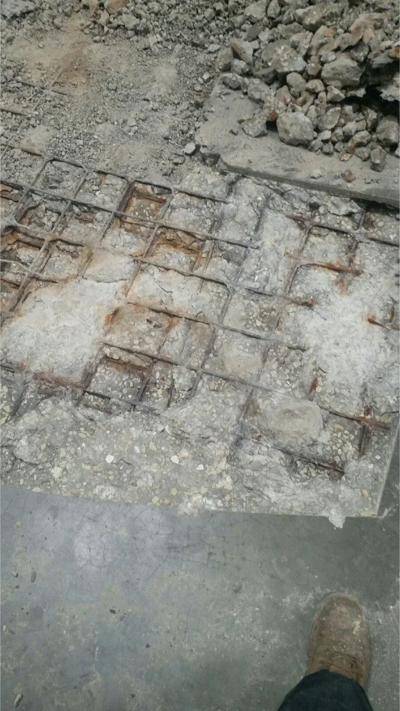 Close-up of demolished slab showing exposed rebar grid and fractured concrete; a worker’s boot is visible at the edge of frame.