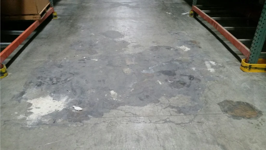 Worn warehouse aisle with patched, spalled, and stained concrete between pallet racking.