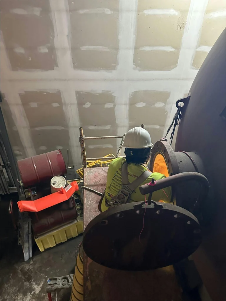 Worker in hard hat and harness on a scaffold at a tank manway, preparing for confined-space entry; drums and ventilation hose nearby.