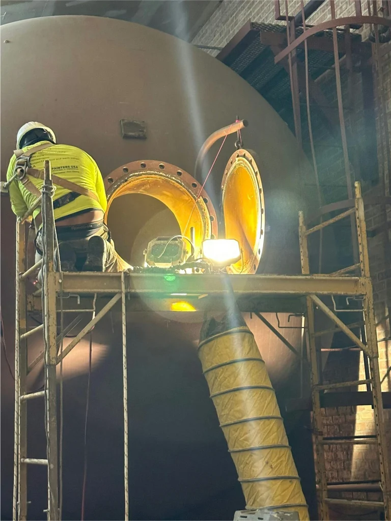 Worker on scaffold leaning into an illuminated tank manway with ventilation hose for confined-space maintenance.