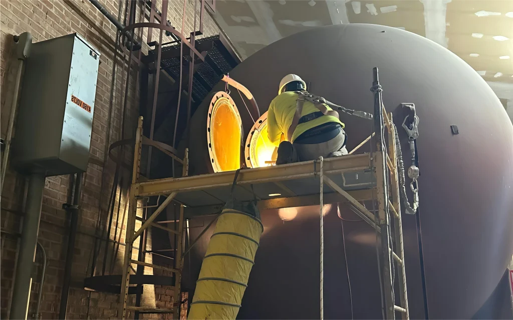 Technician on scaffold at an open manway of a large horizontal vessel; yellow ventilation duct attached.