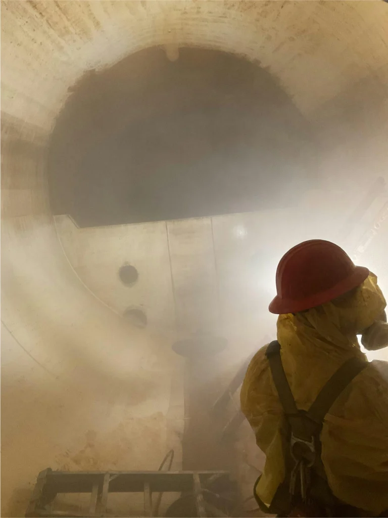 Technician in protective suit, respirator, and hard hat inside a mist-filled tank during confined-space cleaning.