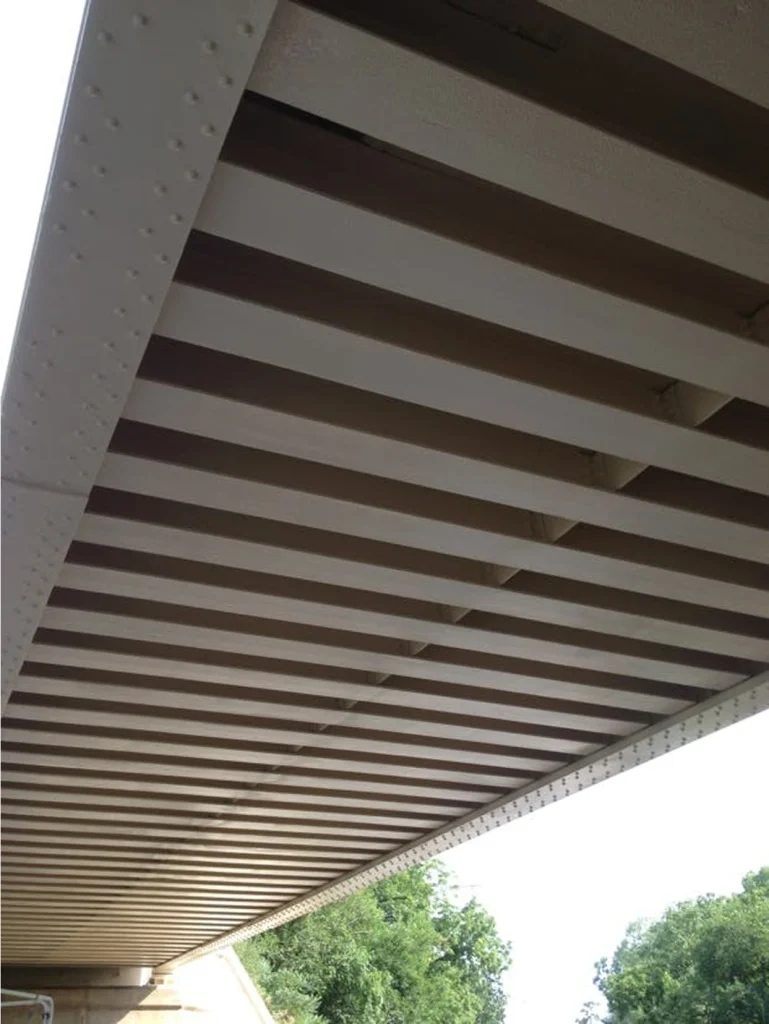 Underside of a steel bridge with evenly coated light-colored beams, showing a clean, uniform finish along the span.