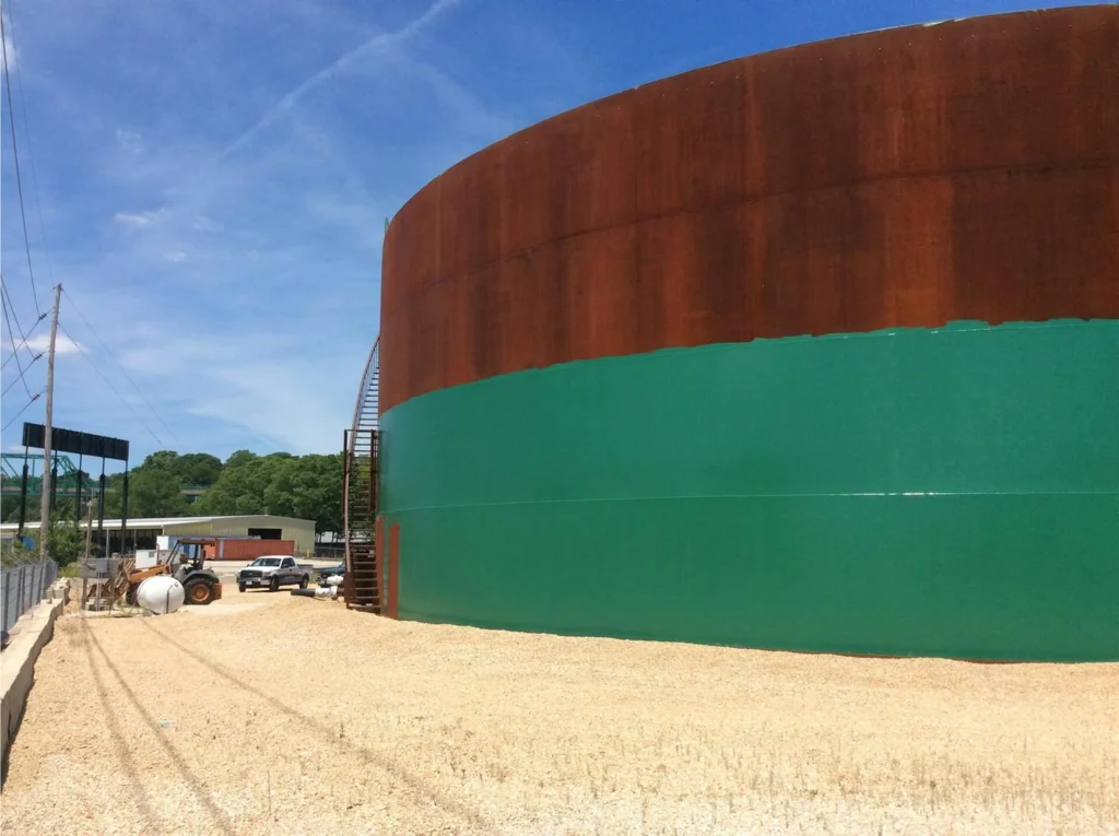 Massive round storage tank mid-project, lower half freshly painted green and upper half still rusted steel.