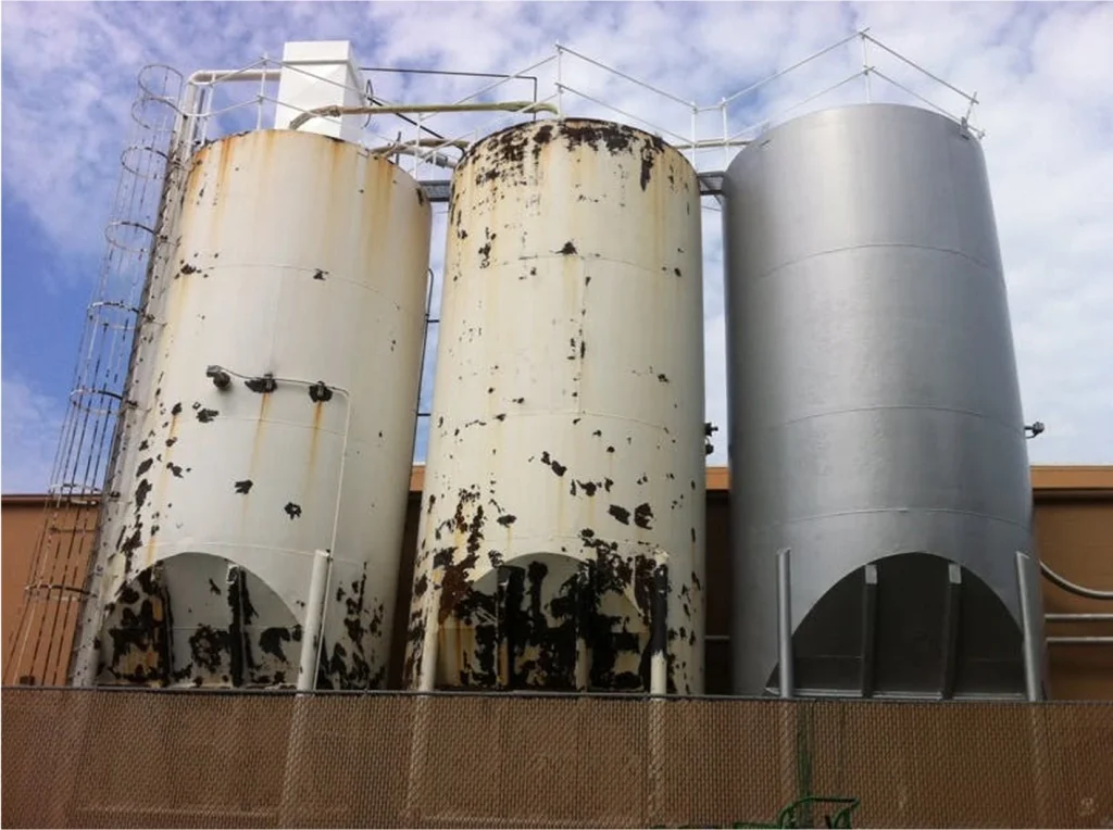 Three vertical silos—two with severe coating failure and rust, next to one newly recoated smooth gray.