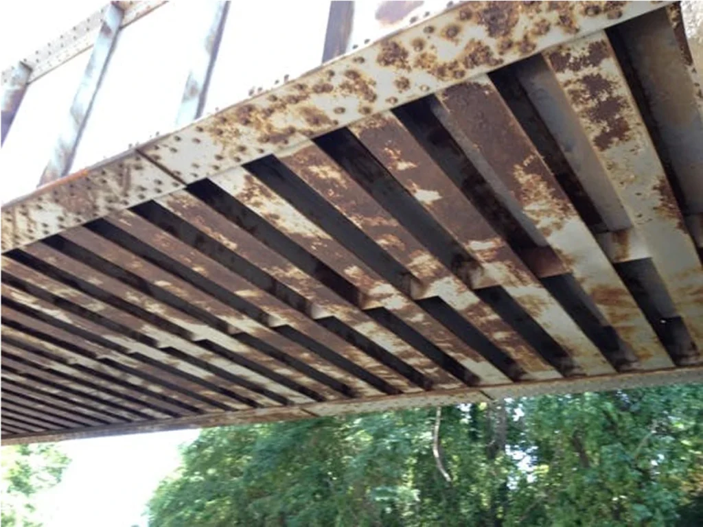 Underside of a bridge before treatment, with heavy rust and flaking paint on the steel stringers.