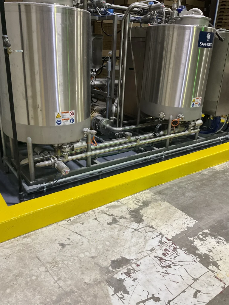 Two stainless-steel processing tanks with pipes, valves, and pumps sit behind a bright yellow safety curb on a worn concrete floor; warning labels are visible.