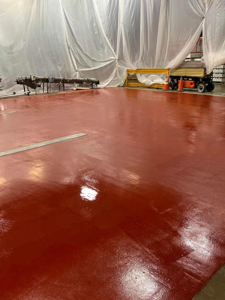 Freshly coated red epoxy floor in an industrial area, still glossy and wet, with plastic containment curtains, traffic cones, a small conveyor, and a yellow safety barrier along the perimeter.