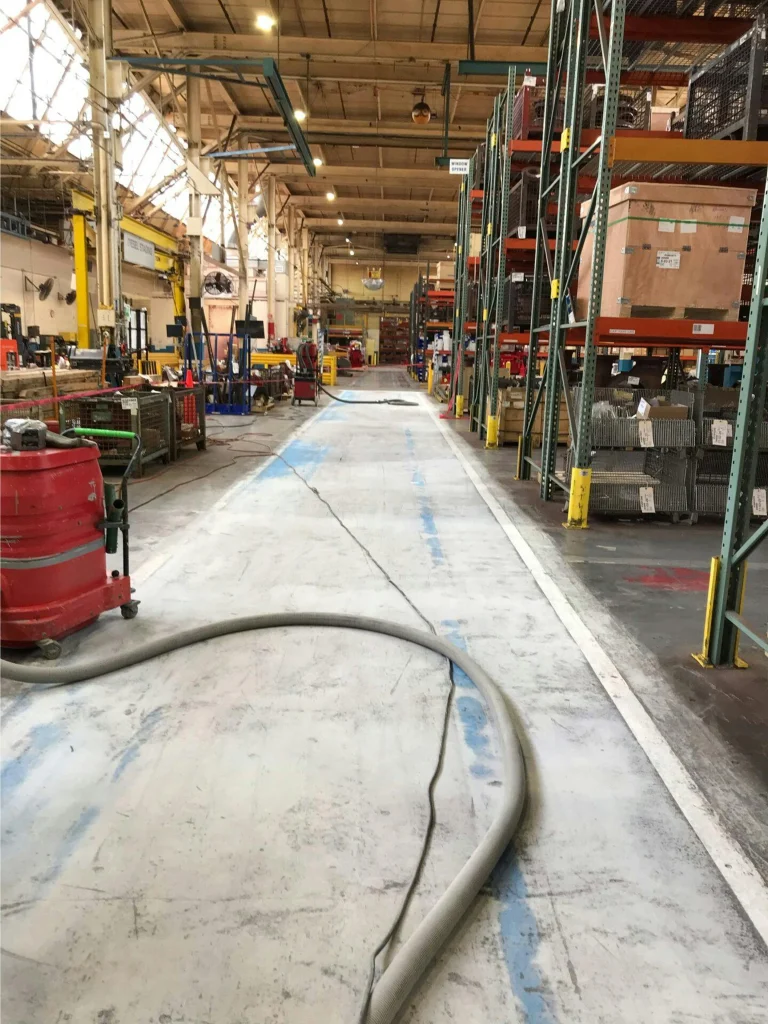 Factory aisle before refinishing: worn concrete with faded blue lines and hoses crossing the walkway.