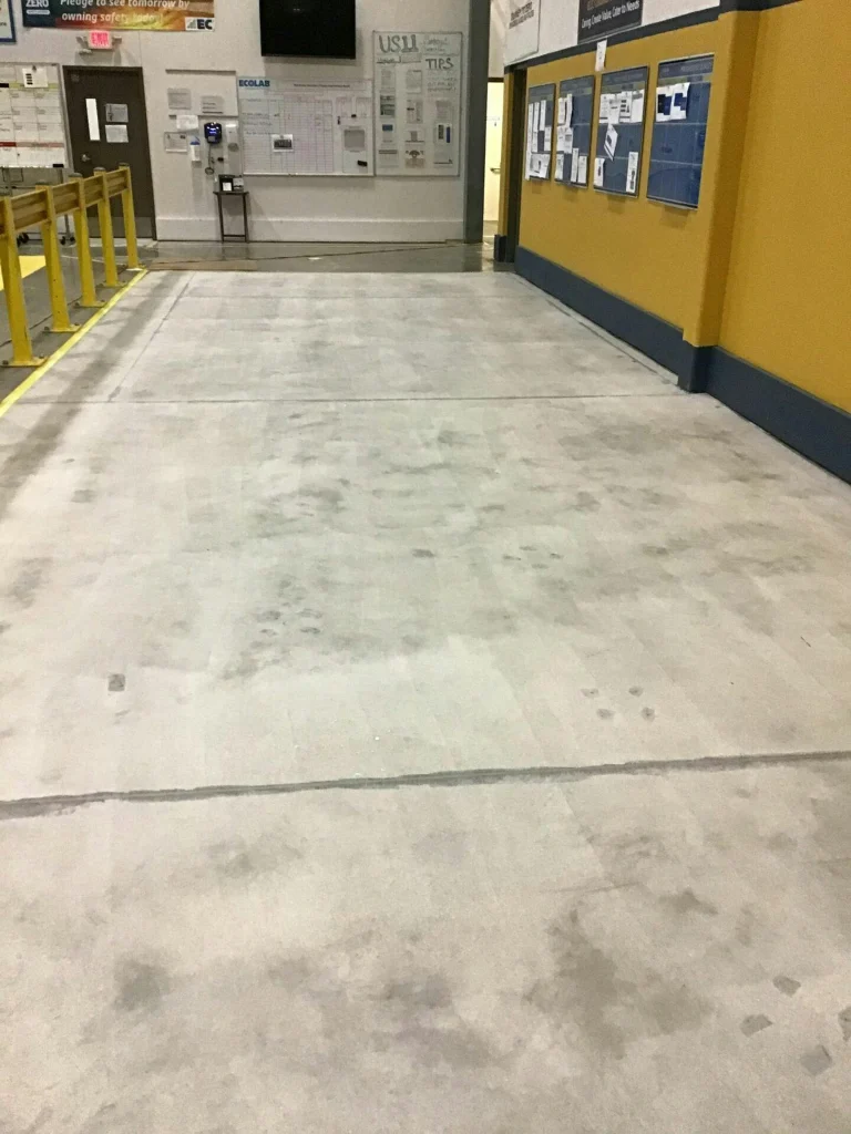 Hallway before coating: ground, bare concrete with saw-cut joint and footprints by yellow rail.