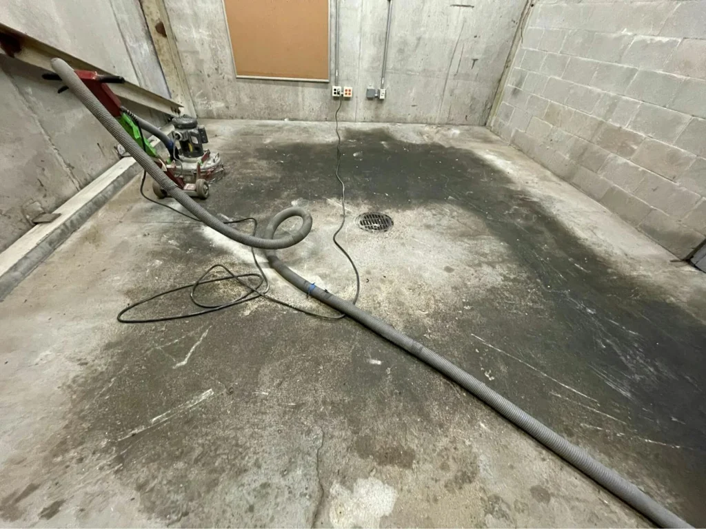 Utility room before: stained, scarred concrete with grinder and vacuum hose near floor drain.