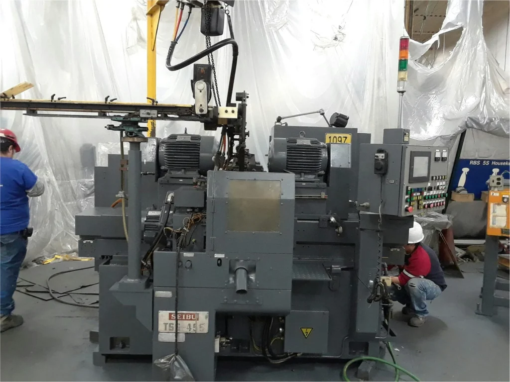 Gray production machine mid-reassembly in a plastic-contained work zone while a worker crouches to service the controls.