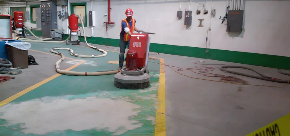 Worker using a large floor grinder to remove old coating in a factory aisle; dust hoses and caution tape line the work zone.