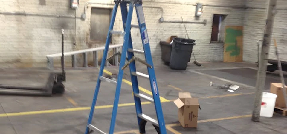 Warehouse corner with a blue step ladder on marked concrete, scattered tools and boxes, and painted brick walls in the background.