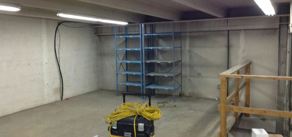 Small storage room with bare concrete floor, fluorescent lights, blue metal shelving, and a coiled extension cord on equipment.