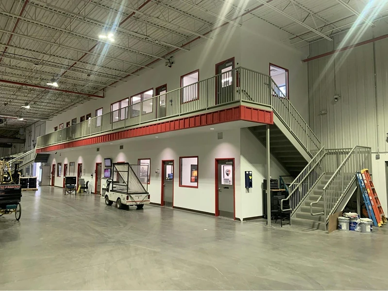 Wide view of a bright industrial warehouse interior with a white, two-story office mezzanine. Freshly painted white walls with red accents contrast with gray railings and stairs under a high, exposed ceiling.