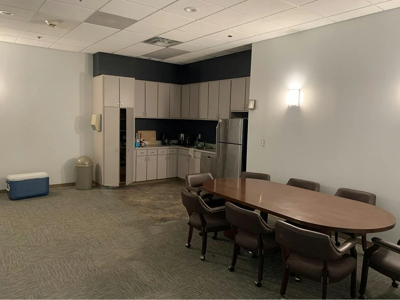 Staff kitchen and meeting space with gray cabinetry, stainless refrigerator, and a long wood conference table. Soft gray walls and ceiling lights create a neat, updated appearance.