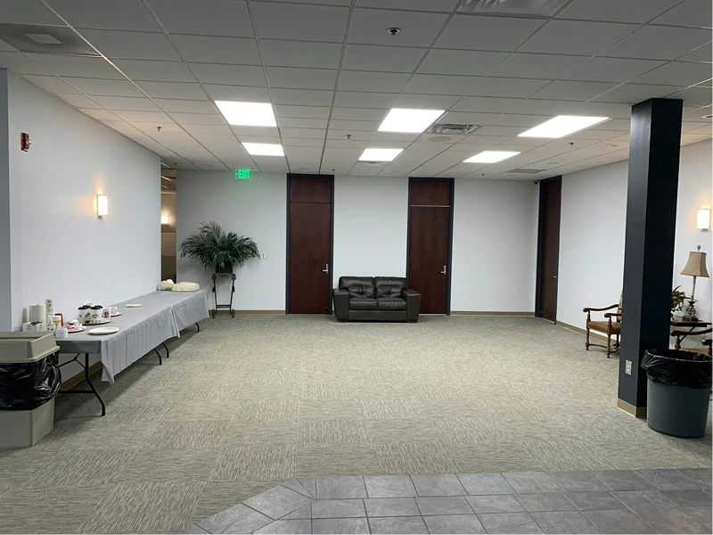 Large open break area or corridor with white walls, dark wood doors, and acoustic ceiling panels. Simple furnishings and bright, even lighting emphasize the fresh paint and spacious layout.