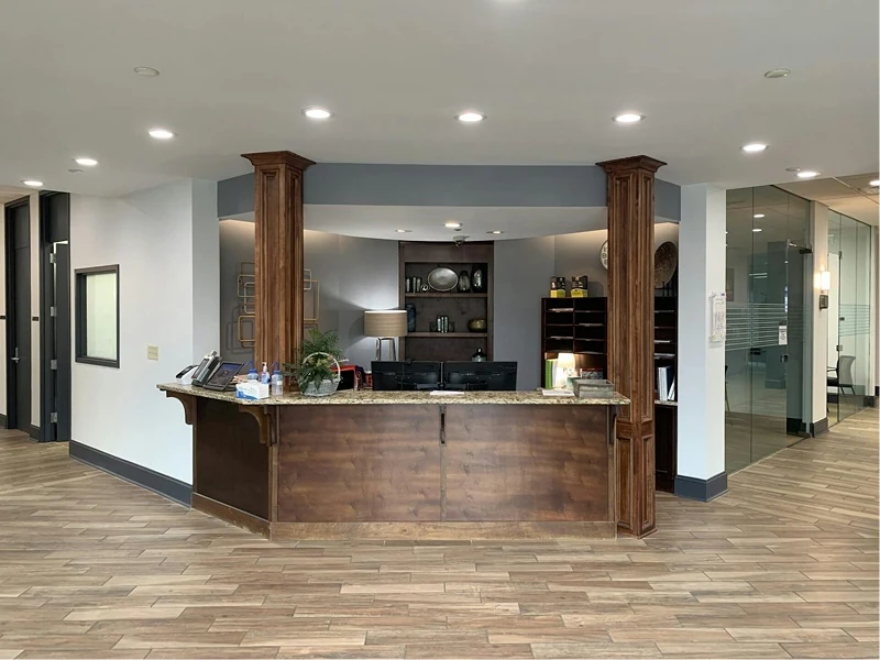 Reception desk with rich wood finishes centered under recessed lighting, surrounded by freshly painted light-gray walls and glass-walled offices. Decorative shelving and artwork complete the professional lobby.