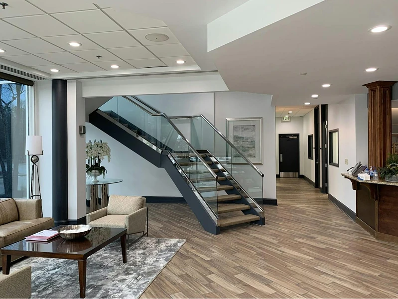 Modern office lobby featuring a glass-railed staircase, wood-look flooring, and seating area with sofas and side tables. Walls are painted in light grays with dark trim for a clean, contemporary look.