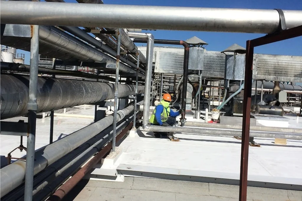 Worker in PPE on a rooftop performing maintenance among large utility pipes and HVAC equipment.