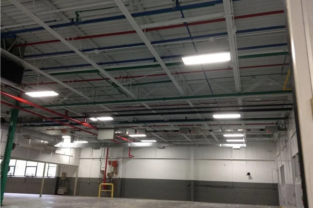 Warehouse ceiling with color-coded pipe runs in red, blue, green, and black against white trusses and lights.