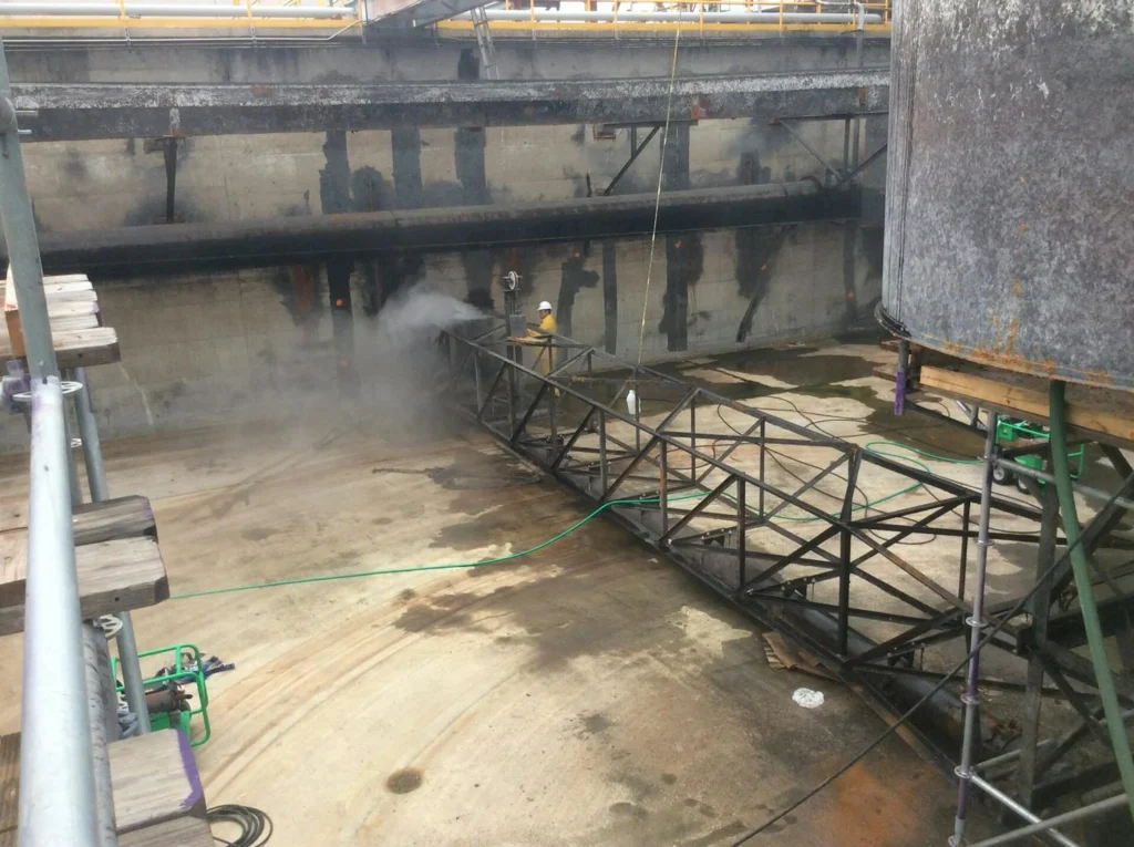 Worker in protective gear power-washing the interior wall of a large concrete basin, with mist rising around a long metal truss walkway.