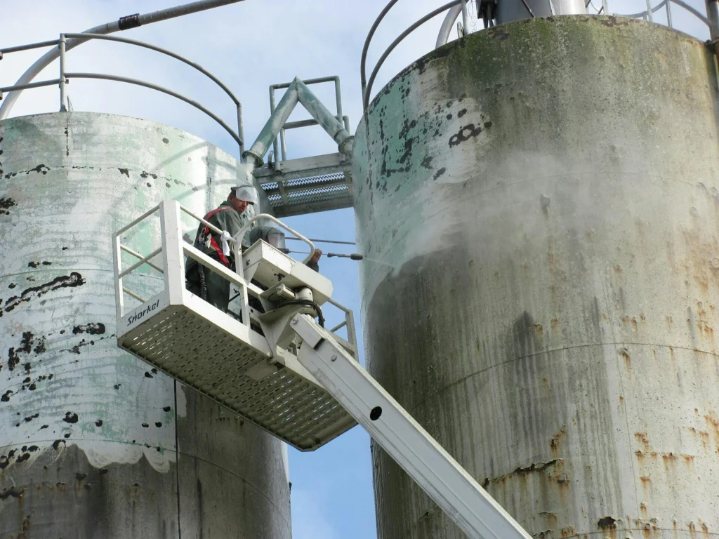 Two weathered industrial silos being pressure-washed from a white boom lift, exposing cleaner concrete where paint and rust are removed.