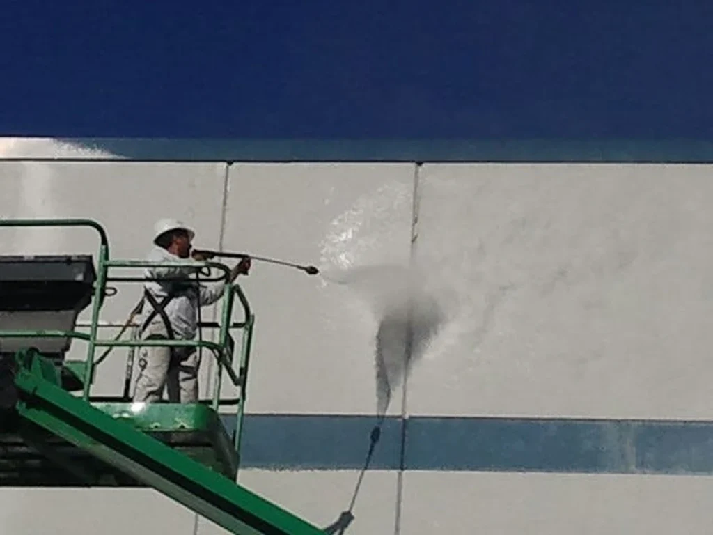 Worker on a green aerial lift pressure-washing a building façade, a bright spray plume revealing a cleaner patch on the white wall.