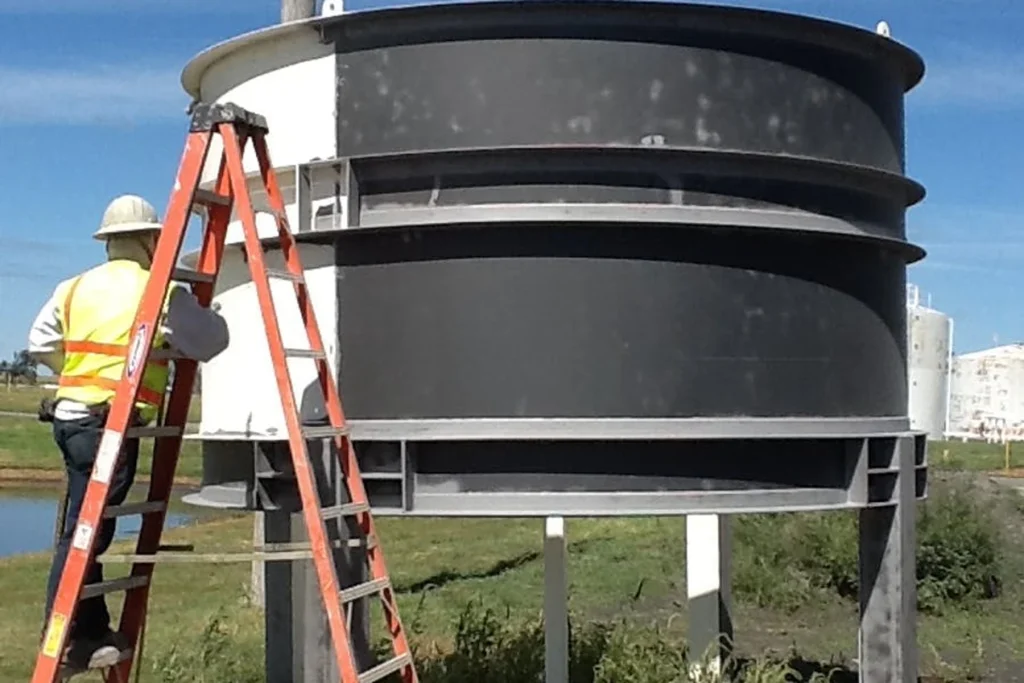 Crew member on a ladder cleaning and recoating a small elevated cylindrical vessel; the shell shows fresh gray coating next to uncoated white areas.