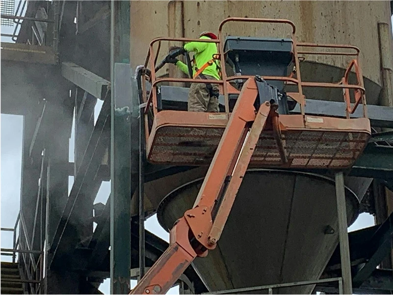 Worker in hi-vis gear on a boom lift wet-vapor blasting rusted steel around a large cone-shaped hopper at an industrial site.