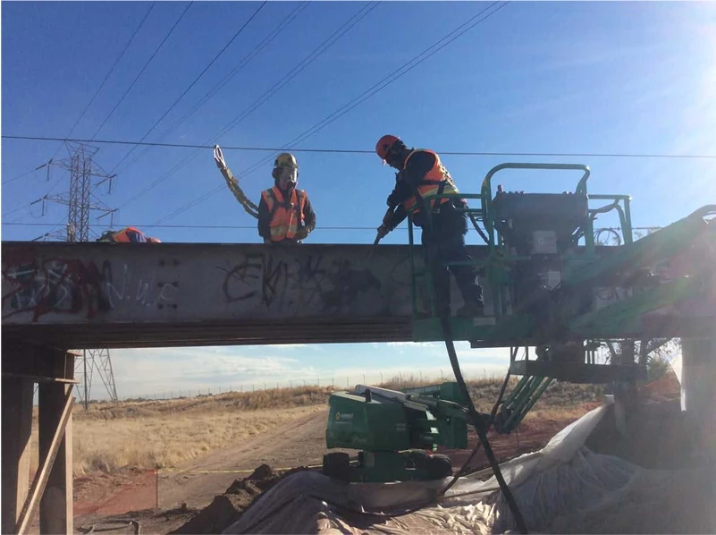 Two workers in fall protection clean graffiti-marked steel on a low bridge from a green boom lift with ground containment below.