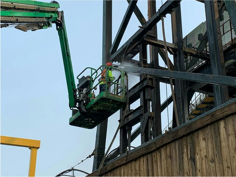 Operator in a green basket directs a wet-vapor blasting stream onto a cross brace of a weathered steel tower above a wooden enclosure.