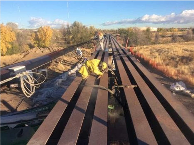 Crew in protective suits wet-vapor blasting long steel bridge girders outdoors, with hoses and containment laid along the span.