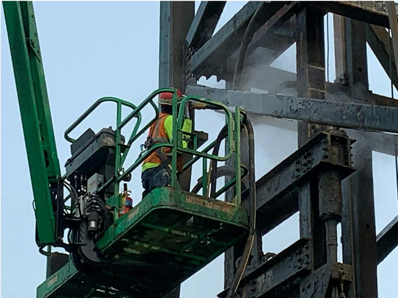 Technician in a green lift uses wet-vapor blasting to clean corroded steel beams on a tall structure, mist visible at the point of cleaning.