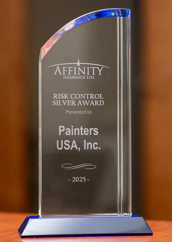 A tall, curved crystal award with blue accents on the top edge and base stands on a wooden surface against a soft brown background. The front is engraved with "Affinity Insurance Ltd." above the heading "Risk Control Silver Award," with text noting it is presented to Painters USA, Inc., and the year "2025" centered near the bottom.