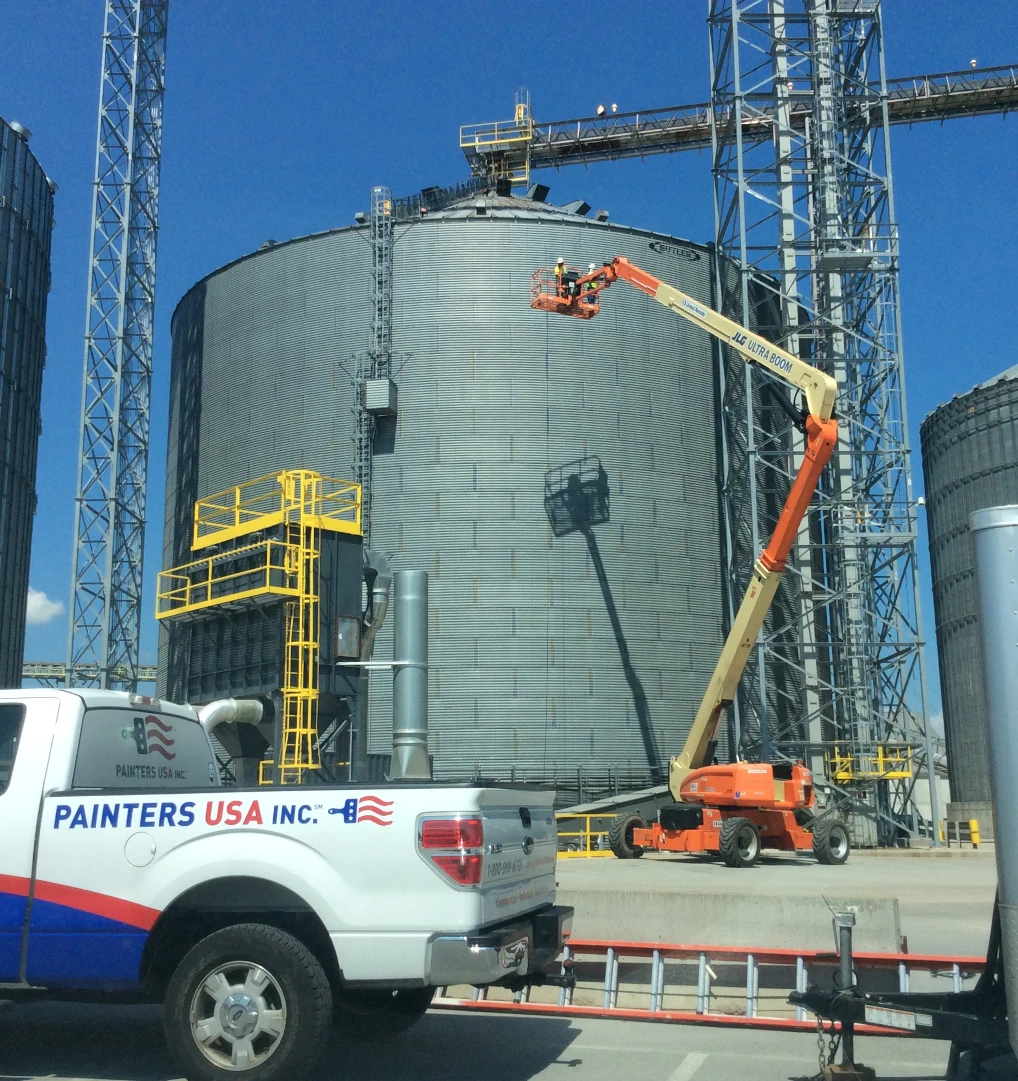 Large gray industrial storage tank at an industrial site with two painters in safety gear on an orange boom lift, and a white Painters USA pickup truck parked in the foreground.