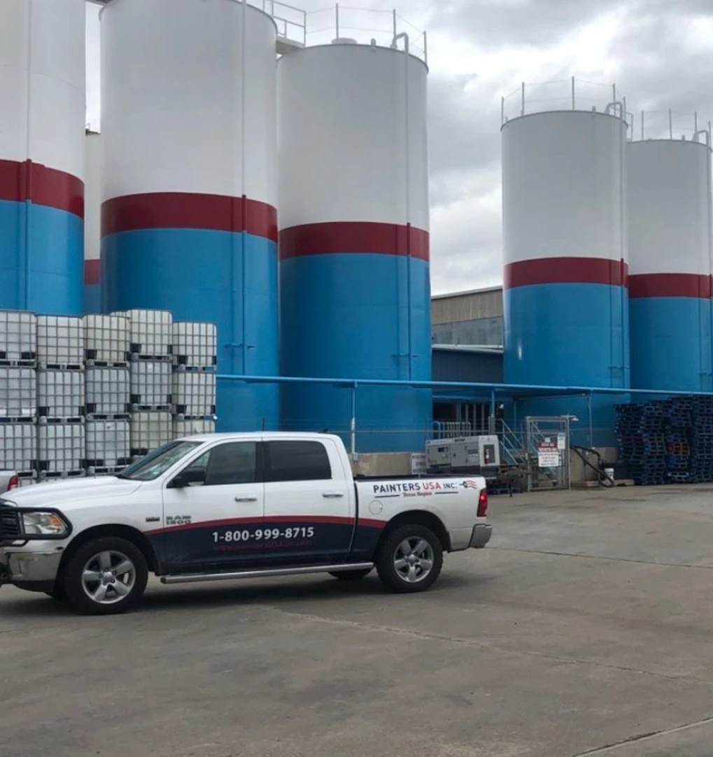 Row of tall cylindrical storage silos painted white with blue bases and a red band, with a white Painters USA pickup truck parked on the concrete surface in front.