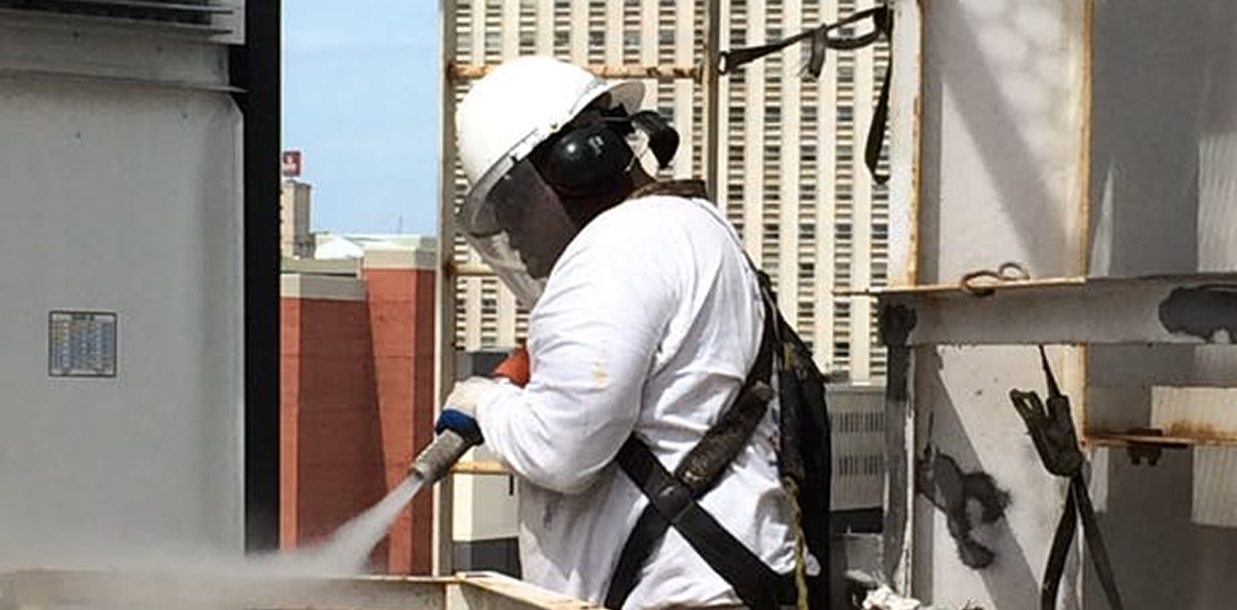 Worker wearing a white hard hat, face shield, and hearing protection uses a high-pressure hose to clean equipment on an outdoor industrial platform; a safety harness is visible and city buildings are in the background.