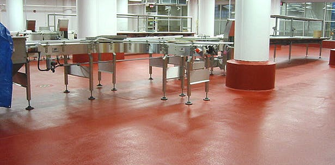 Smooth red industrial flooring in a clean production area with stainless steel conveyor equipment; white support columns with coved bases and bright overhead lighting.