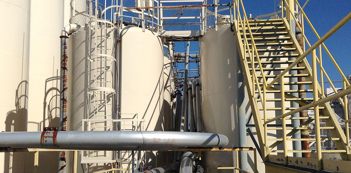 Two large vertical storage tanks and surrounding structures on an industrial site; metal stairs and caged ladders rise alongside the tanks, with interconnected pipes—some showing rust—under a clear blue sky.