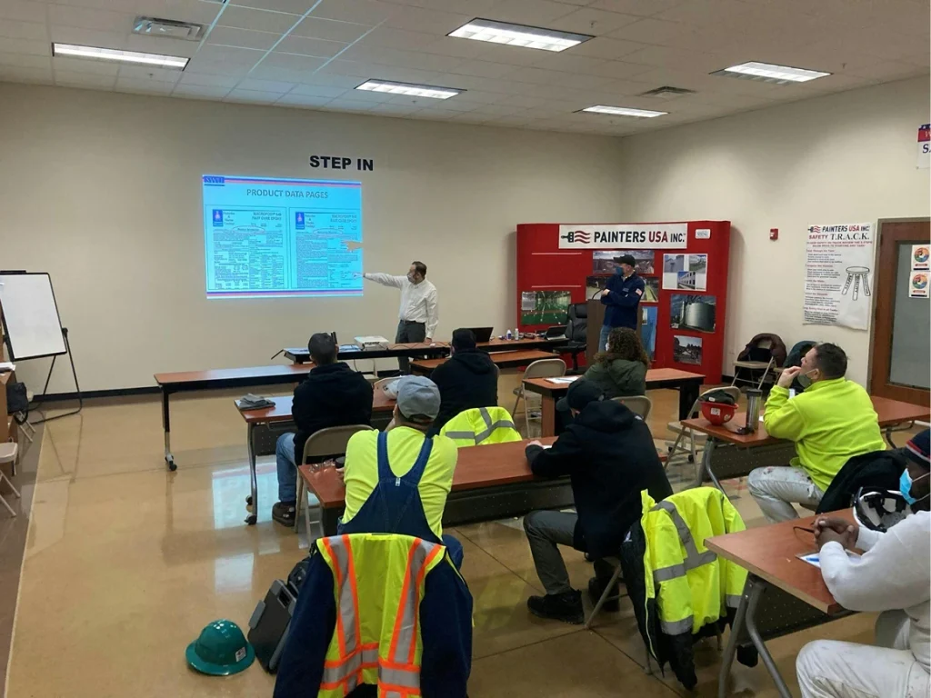 Classroom safety training: instructor points at a projected slide while workers in high-visibility gear listen at spaced desks.