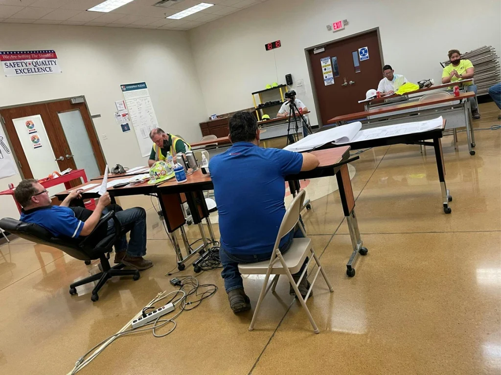 Plan review session: workers seated at tables discuss drawings in a training room with safety posters and visible cables on the floor.
