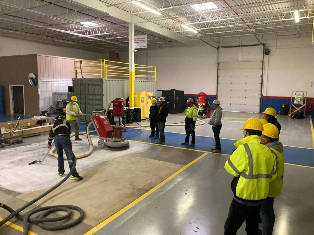 Large shop floor demonstration: crew vacuums and grinds a taped-off concrete section while others observe in PPE.